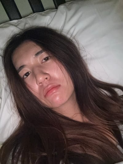A close-up, high-angle portrait shows a young adult female, appearing to be of East Asian descent, lying down indoors. Her long, dark brown hair frames her face, falling across her shoulders and chest. She has a fair complexion with a slight reddish tone on her cheeks and chin, and her lips are full, slightly parted, and glossy. Her dark eyes are open, looking directly towards the camera, and her expression is neutral, possibly appearing drowsy or subtly pouting. She is resting on a white pillow, which fills the right side and upper portions of the background. A dark, vertical element, consistent with a bed frame or headboard, is partially visible along the left edge of the frame. A small portion of a grey garment is visible on her lower right side. The lighting is soft and diffuse. No visual information within the image permits the identification of its location in the city of Le Havre, France, or any other specific geographical area.