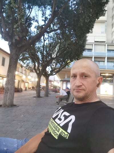 In Follonica, Italy, on a cobblestone square lined with trees and shops, a man is seated on a bench. He has a shaved head and is wearing a black t-shirt with a white NASA logo and yellow text. He is also wearing blue jeans and a silver necklace. In the background, other people can be seen walking and sitting, and a modern building stands tall. The lighting suggests it is either early morning or late afternoon.