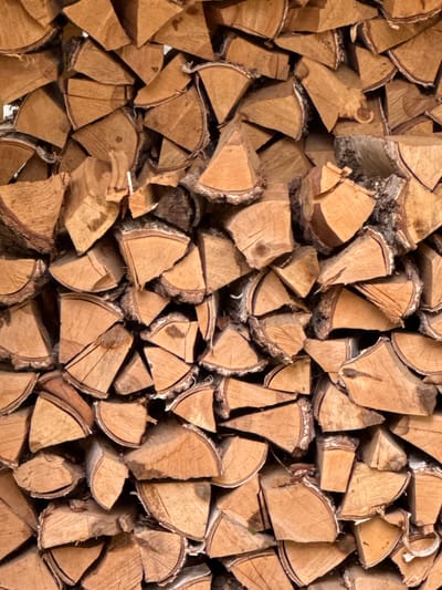 The image shows a tightly packed stack of chopped firewood. The wood pieces are mostly of similar size and shape, cut into wedges with rough, textured bark still attached to some of the outer edges. The grain of the wood is clearly visible on the cut surfaces, appearing in shades of light brown and tan with darker lines. The arrangement is dense, with pieces interlocking to form a solid wall of fuel. There are no people or animals visible. The background is entirely filled with the woodpile, suggesting it's either a close-up shot or a very large stack. The lighting is even, highlighting the texture and natural colors of the wood. No specific location in Газета Труд Снт, Russia can be determined from the image alone.