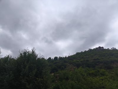 The image depicts a house situated on a lush, tree-covered hillside under a cloudy sky. The house is dark in color and appears to be multi-story with a prominent roofline. Below the house, the hillside is densely vegetated with various shades of green trees and foliage. A visible structure, possibly a telephone pole or light post, is present on the slope. In the foreground, the image is partially obscured by dark green trees, suggesting a roadside perspective. The overall scene conveys a natural, somewhat remote setting.
