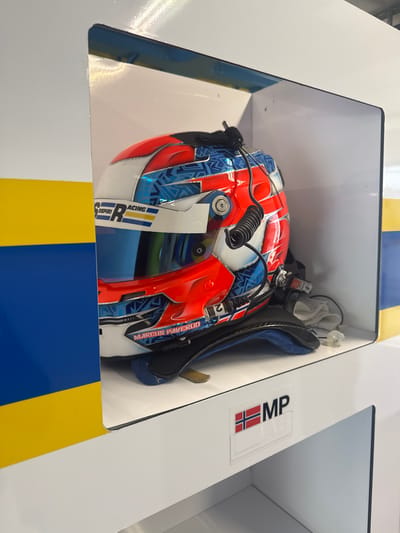 A racing helmet sits within a white alcove, decorated with a yellow and blue horizontal stripe across the top and bottom. The helmet is predominantly red, white, and blue, featuring a design reminiscent of the Norwegian flag. A visor is attached to the helmet, and a label reads "Marcus Paverud." A white cloth and black apparatus are positioned behind the helmet. An "MP" sticker, along with a small Norwegian flag, are on the right side of the alcove. This is located at Vallelunga Circuit Campagnano di Roma, in Campagnano di Roma, Italy.