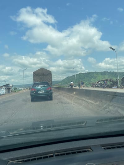 A green Toyota Avensis is seen from behind, driving on a highway. The car has a tinted rear window and a license plate that appears to read "SUG 44UD". To the left of the car, a large truck with a brown tarp covering is also on the highway. The road is paved and shows signs of wear and tear. On the right side of the highway, there is a concrete barrier. Beyond the barrier, there are several makeshift stalls and a group of people. Some are standing, while others are seated or gathered around motorcycles. The background features lush green hills under a bright blue sky with scattered white clouds. Streetlights are visible along the highway. The image appears to be taken from inside another vehicle, looking forward.