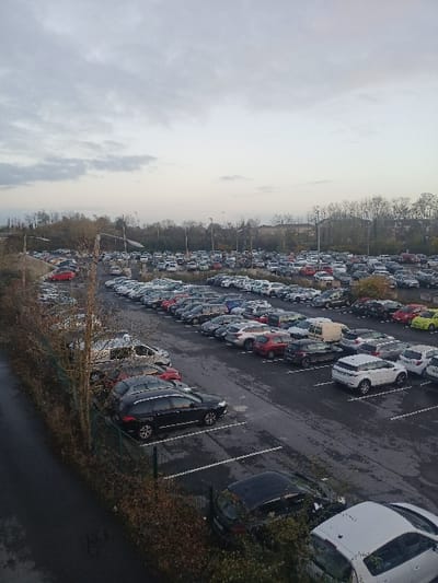 This image captures a large outdoor car park filled with numerous vehicles, viewed from an elevated perspective. The photograph is rotated 90 degrees clockwise, presenting the ground and parked cars diagonally across the frame, with the sky occupying the left side.
Hundreds of cars, primarily compact and mid-sized sedans and hatchbacks common in Europe, are arranged in dense, orderly rows within marked parking spaces. Various colors are present, including white, black, grey, red, and blue. Specific models like a white Peugeot 3008 SUV and a dark grey Renault Clio are discernible among the parked vehicles. The asphalt pavement appears damp and dark in sections, suggesting recent rain or wet conditions.
Along the right side of the car park, rows of vehicles extend towards a distant treeline, which features a mix of bare deciduous trees and some evergreen foliage. Several utility poles or light poles are scattered throughout the parking area and along the treeline.
The sky is heavily overcast with uniform grey clouds, indicating a cloudy daytime. The overall lighting is soft and diffused. No people are visible, and there is no apparent activity other than the static presence of parked cars. The extensive number of vehicles suggests a commercial facility, such as a car dealership's inventory, a large rental car depot, or a vehicle storage and distribution center in Béthune, France. There is no visible text in the image.