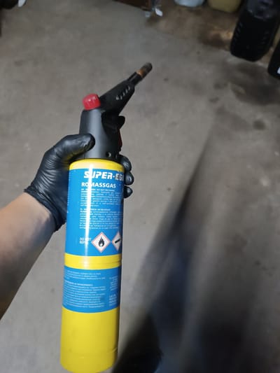 A gloved hand holds a gas torch in a seemingly indoor, utilitarian setting, likely a garage or workshop. The primary subject is a person's left arm and hand, clad in a black, possibly nitrile or latex, glove. The hand firmly grasps the black handle of a gas torch, which is attached to a yellow and blue gas cylinder. A red knob is visible on the torch's assembly. The background consists of a grey concrete floor, with blurry, dark objects that could be tires or containers in the upper right. A shadow extends diagonally across the floor from the lower left towards the upper right.
The visible text on the gas cylinder identifies the brand as "SUPER-EGI ROMASSGAS". Prominent warnings in multiple languages are displayed. In English, it states: "WARNING: DO NOT RECHARGE. DANGER: extremely flammable gas. Contains gas under pressure, may explode if heated. Keep away from heat/sparks/open flames/hot surfaces. No smoking. In case of fire: Do not extinguish, unless leak can be stopped. Eliminate ignition sources if safe to do so. Do not expose to sunlight. Store in a well-ventilated place, a well-covered place." The label also clearly states "DO NOT REFILL." The gas type is identified as "PROPYLENE". The general tone of the labels is cautionary, emphasizing safety due to the highly flammable nature of the gas.