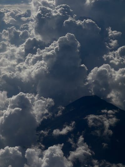The image shows a view of the sky with a large formation of clouds. The clouds are predominantly white and gray, with varying densities and textures. Some areas of the sky are dark, suggesting an overcast or stormy condition. A dark, triangular shape is also present, possibly a mountain. The photo appears to be taken from an elevated position. There is no evidence to pinpoint the location in Taoyuan City, Taiwan. No people, subjects, objects, actions, or interactions are visible in the image.