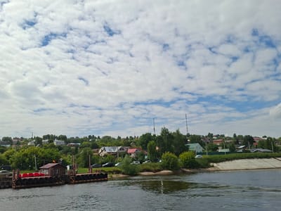 The image depicts a tranquil scene on a river in Verkhniy Uslon, Russia. In the foreground, a ferry dock is visible, constructed with dark, tire-like buffers along its side. A small, brown building with a red roof sits on the dock, and several rows of red seating are arranged behind it. The water in the river appears calm with slight ripples.
The middle ground showcases a verdant hillside densely covered with trees and houses. The architecture of the houses varies, with some featuring red roofs and others in lighter colors. A few cars are parked along the roadside that winds through the settlement. In the distance, two tall radio towers are prominently visible on the horizon. The sky is partly cloudy, with a mix of white cumulus clouds scattered across a blue expanse. The overall impression is one of a peaceful, semi-rural setting.