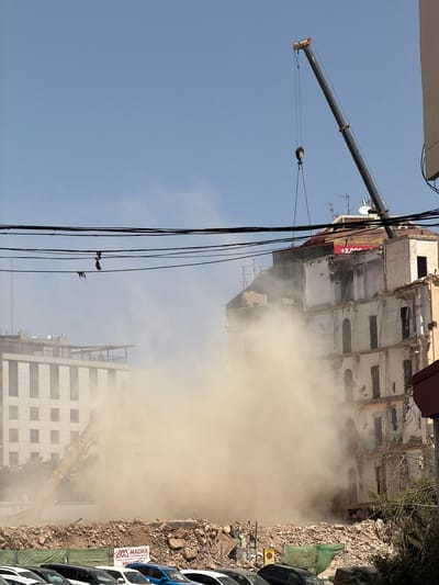 In the city of Unknown, Unknown, a large building is undergoing demolition, with debris and dust obscuring much of the scene. A tall crane with a dark boom extends diagonally across the sky from the right side of the frame, its hook and chains hanging downwards. In the background, a lighter-colored, multi-story building with many windows is partially visible through the haze, its top section appearing to be under construction or renovation. To the left, a yellow excavator is actively working, contributing to the dust cloud. Power lines are strung across the upper portion of the image. The ground in the foreground is covered with rubble and debris from the demolition. Several cars are parked in front of the demolition site, partially obscured by the debris. The sky is a clear, pale blue, with no clouds visible.