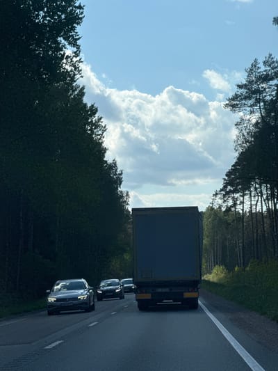A medium-angle shot depicts vehicles traveling on a road bordered by a forest and sky in Virši, Latvia. A large dark truck leads a line of cars, their features and reflections visible in the sunlight. The vehicles move along a paved road marked with a solid white line. The background consists of dense trees on either side of the road and a sky with scattered clouds. The composition focuses on the vehicles and the surrounding natural landscape.