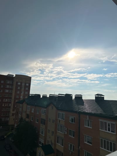 This media file is a vertical, outdoor, daytime photograph taken from a high vantage point, likely a window or balcony, looking up at the sky and the sides of apartment buildings. The sun is prominently visible, creating a lens flare and casting a bright light across the scene. Wispy white clouds are scattered across a blue sky, indicating fair weather.
Two apartment buildings are the main objects in the scene, their brick facades and numerous windows clearly visible. The buildings are modern, with dark, sloped roofs and what appear to be air conditioning units or vents on some of the walls. The architecture suggests a residential area. There are dark green roofs on the buildings closest to the viewer, and a large, dark green tree is visible between the buildings, suggesting a park or green space nearby.
The time of day is clearly daytime, with the sun high in the sky. The lighting suggests it could be morning or afternoon, given the angle of the sun and the bright glare. There are no people visible in the image. The setting is urban, specifically a residential area in Солнечный, Russia. There is no visible text in the image. The overall mood is bright and somewhat serene, dominated by the natural elements of the sun and sky.