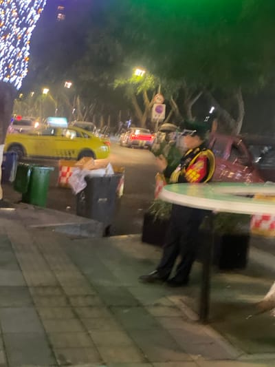 This image depicts a nighttime urban street scene in 天宫殿街道, China.
In the immediate foreground, a tiled sidewalk is visible. On the left side of the sidewalk, a green refuse bin stands next to a dark gray refuse bin, which appears to be overflowing with trash. Behind these bins, red-and-white checkered barriers partially cordon off the street. On the right side of the foreground, a round, light-colored table with a dark base and some decorative greenery is partially visible.
A person is seated or leaning near this table, facing towards the left side of the frame. This individual is wearing a dark uniform, consisting of a peaked cap, what appear to be sunglasses, a dark jacket adorned with prominent red and gold insignia on the chest and shoulders, dark trousers, and dark shoes. The person's hands are clasped, holding a small, indistinct object, possibly a mobile phone.
In the midground, a bright yellow taxi is positioned on the street, facing away from the viewer. Further down the street, other vehicles with illuminated lights are visible in what appears to be traffic or parked positions. The street is lined with trees, whose foliage is illuminated by several streetlights positioned along the road. A circular traffic sign is discernible on the right side of the street. On the far left, a vertical string of numerous small, bright lights, possibly decorative, extends upwards. The scene is illuminated primarily by artificial lights, indicating nighttime conditions.