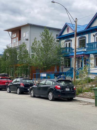 The image depicts a street scene in Whitehorse, Canada, showcasing a row of buildings and parked vehicles. On the right side of the frame, a historic-looking building with white siding, blue trim, and a bright blue roof stands prominently. Decorative blue railings adorn its balconies and staircases. To the left of this building is a more modern structure with corrugated metal siding and a flat roof, featuring large windows and a balcony.
Several cars are parked along the street. In the foreground, a black Subaru hatchback is parked facing away from the camera. To its left, a black Toyota hatchback is also parked. Further down the street, a red SUV is visible. The street itself is paved asphalt, and a sidewalk runs along the right side of the frame, bordered by a grassy area with some vegetation. A green garbage bin is situated on the sidewalk. A street lamp with a curved arm is visible overhead, attached to a wooden utility pole. The sky is overcast with cloudy conditions. There are no people visible in the image. The scene appears to be a commercial or residential area within the city.