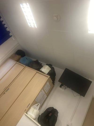 The image depicts a room with a large wooden cabinet occupying the left side. On top of the cabinet, several trash bags of varying sizes and colors are piled, along with a brown cardboard box. To the right of the cabinet, a black television screen is mounted on a white wall. Below the television, a black backpack is placed on the floor. In the lower right corner, a white bag filled with white objects is visible. The room features a white tiled ceiling with a fluorescent light fixture and a smoke detector. The scene is devoid of any individuals. The exact location within Қосшағыл ауылдық округі, Kazakhstan, cannot be determined from the image alone.