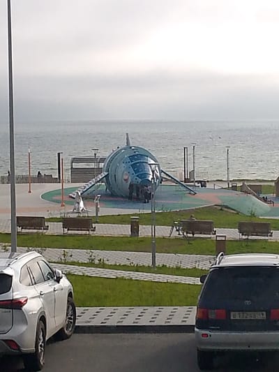 The image shows a playground designed to look like a rocket or spaceship, situated near a body of water, likely a sea or large lake, under an overcast sky. The playground structure is large and metallic blue, with windows and a ramp leading up to it. Several people are visible near the structure, some entering or exiting. In the foreground, two cars are parked: a white SUV on the left and a dark minivan on the right. The minivan has a license plate that appears to read "B 132 AA 59". The area around the playground is paved with patterned tiles and grass, with benches and trash receptacles present. In the background, a railing separates the paved area from the water, with a few distant figures visible on the other side of the railing. The overall setting suggests an outdoor recreational area in a coastal or lakeside city. The license plate suggests the location is in Russia. Given the context of a prominent spaceship-themed playground near the sea in Russia, this scene is likely located at the "Navigator" Space-themed Playground in Magadan, Russia, a well-known local attraction.