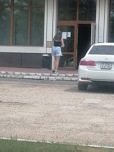 A young woman with long, dark hair is standing on a paved area in front of a building with large windows and a glass door. She is wearing a black tank top, a blue denim skirt, and white sneakers. She is facing away from the camera and appears to be holding something in her left hand. A white sedan is parked to the right of the woman, partially obscuring the building's entrance. The license plate of the car reads "P 310 XK 125". The background shows a grassy area and a street. The overall impression is a casual outdoor scene. This image was captured in the city of Lesozavodsk, Russia.