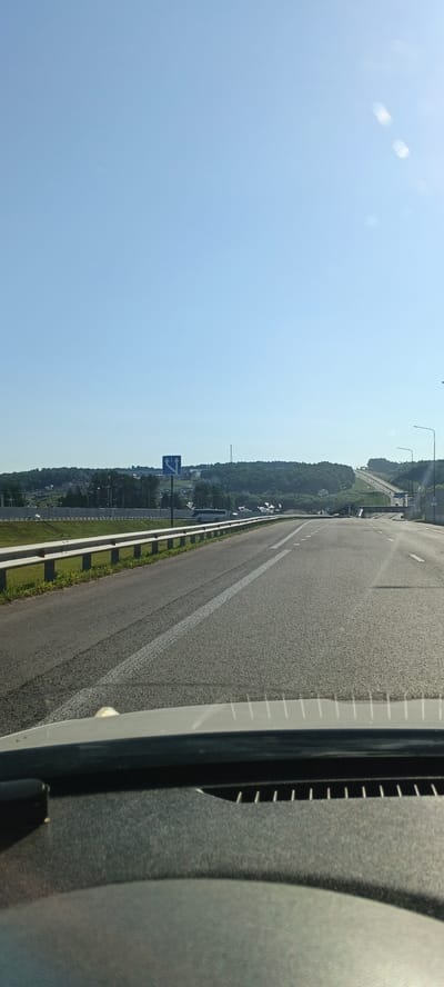The image is taken from the perspective of a car dashboard, looking out at a highway. The highway stretches into the distance, curving gently to the right. The sky is clear and blue with a few faint glints of light. On the left side of the highway, there is a grassy embankment with a metal guardrail. Beyond the embankment, there are trees and buildings, suggesting the highway is near a populated area. A blue traffic sign with white arrows indicating lane changes is visible. On the right side of the highway, there are streetlights and the road continues to ascend. The foreground shows the textured surface of the car's dashboard. The overall scene depicts a travel or journey on a highway under a clear sky, possibly in or around the city of Staroe Koshchakovo, Russia, as suggested by the prompt. There are no visible people, and the focus is on the road and the surrounding landscape.
