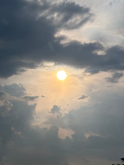 The image captures a dramatic sky with the sun partially obscured by dark, textured clouds. The sun, a bright, intense orb, casts a warm, golden light that diffuses through thinner patches of clouds, creating a hazy glow. The clouds themselves are varied in form and color, ranging from deep grays and blues to lighter, more wispy formations catching the sunlight. Some clouds appear heavy and dense, suggesting a possibility of rain, while others are translucent, allowing the sun's rays to penetrate and create a luminous effect. The overall impression is one of a dynamic and atmospheric sky, possibly during the late afternoon or early evening as indicated by the warm lighting. There are no people, man-made objects, or discernible location cues like buildings or landmarks. The scene is purely natural, focusing on the interplay of light and shadow within a cloudy sky. The emotional tone evoked is one of awe and perhaps a touch of foreboding due to the contrast between the bright sun and the dark clouds. No text is visible in the image.