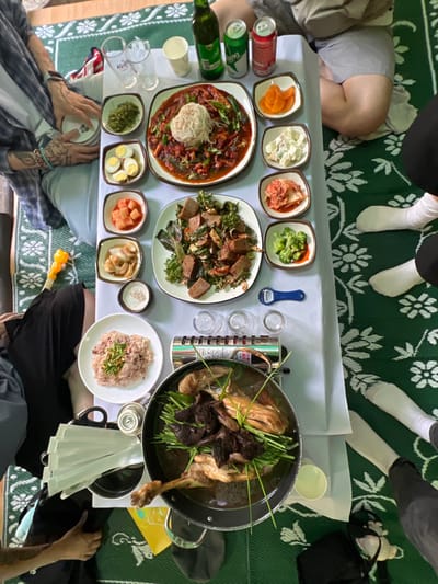 An overhead view depicts a group meal taking place indoors in 강북구, South Korea. Several individuals are seated on a green mat with white floral patterns on the floor around a low table covered with a white disposable tablecloth.
Visible individuals include:
* On the left, a person wearing a plaid shirt over a white t-shirt and light-colored pants, with multiple tattoos on their forearms and wrists, holding an empty white plate.
* In the upper right, a person wearing a light grey top and shorts.
* The legs and feet of at least two other individuals are visible; some wear dark pants and white socks.
The table is densely covered with numerous Korean dishes and beverages:
* **Main Dishes:** A large square platter holds a red-sauced stew, possibly seafood or meat, topped with white somyeon noodles and green onions. Another plate features a green leafy salad with dark brown, rectangular pieces. A large metal pot, situated on a portable gas stove, contains a simmering stew with a whole or partial duck/chicken, dark ingredients (possibly mushrooms or herbs), and green chives. A round plate holds a mound of pinkish-brown rice garnished with green.
* **Side Dishes (Banchan):** Arranged in small square white bowls are various items, including cooked green vegetables, four halved hard-boiled eggs, diced white radish kimchi (kkakdugi), pickled garlic or onions, potato salad, red kimchi, blanched broccoli with red sauce, and orange pickled radish or sweet potato. A small circular dish contains white granules, likely salt or sesame seeds.
* **Beverages:** A green beer bottle (identifiable as Kelly beer by the visible glass logo) is present, alongside two soda cans (one green, one red). Two clear drinking glasses and one white paper cup are on the table. Four small clear shot glasses are positioned near the portable stove.
Additional objects include a blue bottle opener, a stack of white paper napkins or tissues, and a partial view of a yellow plastic object in the bottom-left corner. The portable gas stove is actively heating the stew, suggesting an ongoing meal or preparation.