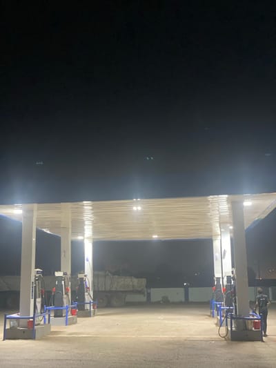 This image depicts a gas station at night in Issele Uku, Nigeria. The scene is illuminated by overhead lights embedded in a white, flat-roofed canopy supported by white pillars with blue trim. Below the canopy, four fuel pump islands are visible, each equipped with multiple dispensers featuring white, blue, red, and black components. In the mid-ground, a dark-colored flatbed truck is parked under the canopy, laden with numerous large, wrapped, rectangular bales secured by netting or ropes. Partially visible to the far left is the rear of a white truck. On the far right, a single individual wearing dark clothing stands upright near a fuel pump island. The ground is paved, and the sky above is uniformly dark.