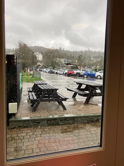 A view through a window shows a rainy day in Bowness-on-Windermere, United Kingdom. Outside, wet cobblestones are visible, along with two dark picnic tables. Cars are parked in a row along the street. The weather appears overcast, with gray skies. Trees are bare, indicating a cold season. There is a building with a sign that is partially obscured by the window frame.