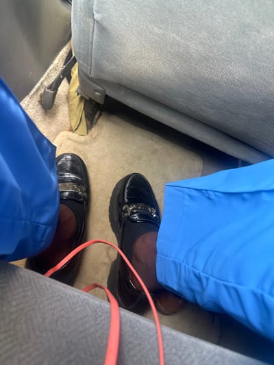 The scene depicts the interior of a vehicle located in Eneka, Nigeria. The perspective is downward, focusing on the lower legs and feet of an individual seated inside. The person has dark skin and is wearing royal blue pants or scrubs, along with dark-colored socks. On their feet are a pair of black, shiny patent leather loafers, each adorned with a silver-toned metal bit across the vamp. These loafers feature thick, ridged soles. In front of the individual, a light gray fabric-covered seat back is visible. The vehicle floor is covered with a beige carpet. To the left, near the floor, a dark-colored metal seat mechanism is visible, alongside a crumpled piece of olive green fabric. A small, dark brown shoe or slipper is partially visible tucked under the right blue pant leg. An orange-red strap or cord arches across the carpet between the individual's feet.