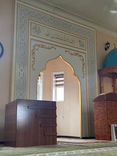 The image depicts the interior of a mosque or Islamic prayer hall, likely in Dehqonbozor, Uzbekistan. The scene is illuminated by natural light, suggesting it is daytime. No people are visible, indicating a quiet moment between prayer times.

The central feature is a richly decorated mihrab, an arched prayer niche that signifies the direction of prayer (qibla). The mihrab's walls are adorned with intricate Islamic geometric patterns in light blue/gray and gold, featuring interconnected star shapes and arabesque motifs. Prominently displayed within the mihrab, in elegant gold Arabic calligraphy, is the Shahada: "لاَ إِلَهَ إِلاَّ اللَّهُ مُحَمَّدٌ رَسُولُ اللَّهِ" (Lā ʾilāha ʾillā llāhu Muḥammadun rasūlu llāhi), meaning "There is no deity but God, Muhammad is the messenger of God." Above this, the word "اللَّهُ" (Allah) is visible, and below it, another calligraphic element, likely related to the Prophet Muhammad.

To the right of the mihrab (left in the rotated image) stands a wooden minbar, a pulpit from which the imam delivers sermons. Further to the right, a dark brown wooden lectern or storage unit with vertical decorative slats is present in the foreground.

In the upper left corner, part of a light blue dome structure is visible, possibly belonging to the mosque's exterior or an internal decorative element. A small, dark wall lamp is also attached near the dome. Windows allowing natural light into the space can be seen through the mihrab's archway, revealing light-colored walls beyond. The floor on the right is covered with a patterned green carpet, indicative of a prayer rug or hall carpeting. The overall ambiance is one of serenity and reverence, typical of a place of worship.
