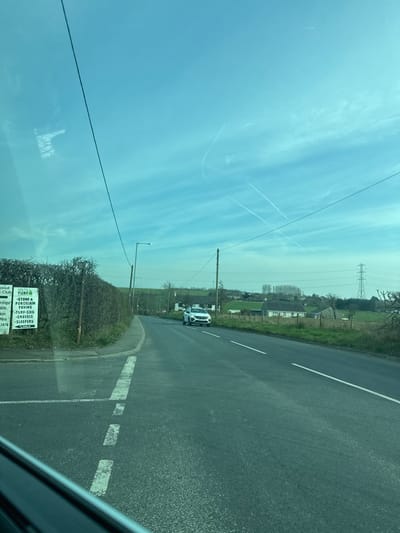 The image displays a two-lane asphalt road under a bright blue sky with scattered cirrus clouds and visible aircraft contrails. The scene is located in a rural or semi-rural area of Bolton, United Kingdom.
In the foreground, the image is framed by the interior of a vehicle, visible in the lower left, with reflections of tree branches on the windshield. The road features white dashed center lines and a solid white line marking the left edge, curving slightly to the left.
On the left side of the road, a dense green hedge runs parallel to the roadway. Behind the hedge, a white sign with green lettering is visible, identifying "Bolton Turf Farm" and listing services including "TURF," "TOP-SOIL," "STONE & PORCELAIN PAVING," "GRAVEL," and "SLEEPERS." A utility pole with overhead wires and a street light pole are positioned along this side.
Ahead on the road, a white SUV, possibly a Hyundai Tucson based on its grille design, is parked or slowly approaching, facing the camera, situated in what appears to be the opposite lane. Its headlights are off.
The background consists of undulating green fields, several scattered trees, and a few detached residential or farm buildings. A prominent large electricity pylon stands on the far right horizon.