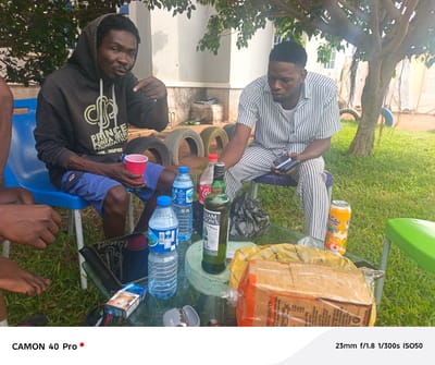 The image captures an outdoor gathering in what appears to be Langtang, Nigeria. Two men are prominently featured, seated on plastic chairs around a low, round glass table. The man on the left wears a dark hoodie with the text "PRINCE PONFA MIR FOUNDATION -RM. INSPIRE" and blue shorts; he's gesturing with a red plastic cup in hand. The man on the right wears a striped button-down shirt and white pants, looking down at a phone or small electronic device in his hands. The table is laden with items: several bottles of water, a bottle of William Lawson's Scotch Whisky, a can of "Vudu" juice, a loaf of bread in plastic packaging, cigarettes, and a black plastic bag. There's also an open lighter. A portion of another person's bare foot and hand are visible in the lower-left corner. Tires are stacked behind the men. The scene is set on a grassy area with trees providing shade; a building is visible in the background. The camera used is a CAMON 40 Pro.