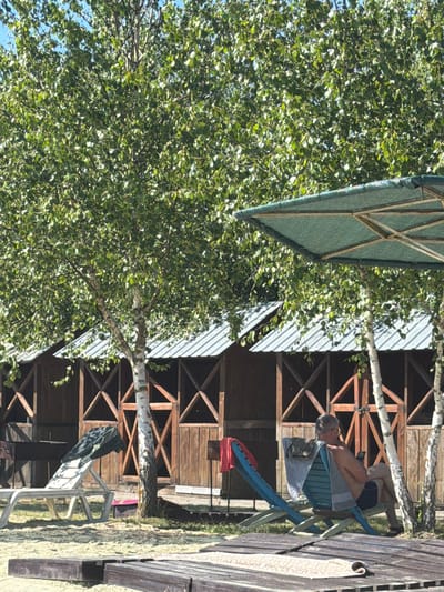 In the city of Verbizh, Ukraine, an outdoor scene unfolds under a bright sky. A man in his late 50s or early 60s is seated in a blue lounge chair, facing away from the viewer. He is wearing dark shorts and appears to be holding a smartphone in his right hand, with his left hand resting on his lap. A light blue towel is draped over the back of the chair, and a darker towel or garment is placed on his shoulders. His hair is gray and he wears glasses.

In the foreground, a white plastic lounge chair is positioned on the sand, with a dark patterned towel or blanket resting on it. A large, green, octagonal umbrella, likely for shade, is partially visible in the upper right portion of the frame.

Behind the chairs and umbrella, a row of wooden cabins with metal roofs is visible. The cabins have open fronts with decorative wooden X-shaped bracing. The surrounding area is sandy, with patches of grass and the trunks of birch trees with distinctive white bark interspersed throughout the scene. The leaves of the trees are lush and green, indicating a warm season. The overall impression is a relaxed, outdoor recreational setting, possibly a beach or a resort area.