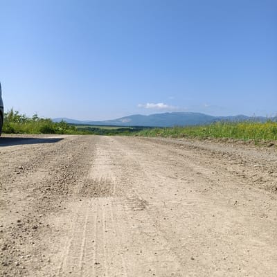 The image depicts a low-angle view of a dirt road stretching into the distance, with the tires of a vehicle visible on the left side of the frame. The road is composed of gravel and dirt, with tire tracks pressed into its surface. To the right of the road, there is a grassy embankment with scattered yellow flowers. Beyond the embankment, rolling hills and mountains form the background under a clear blue sky with a few wispy clouds. The scene suggests a rural or undeveloped area, possibly on the outskirts of Tambovskoe, Russia.
