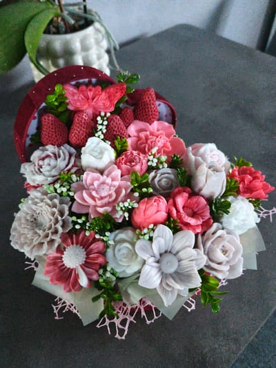 A heart-shaped bouquet filled with handcrafted soap flowers and artificial strawberries is arranged on a dark, textured surface. The bouquet is predominantly in shades of pink, white, and grey, with some red accents from the strawberries and a single red flower. A pink butterfly decoration is visible at the top of the bouquet. In the background, a portion of a potted plant with a green leaf and a white, textured pot is slightly out of focus. The scene is lit from above, casting soft shadows. The location of Brodi, Latvia is not discernible from the image.