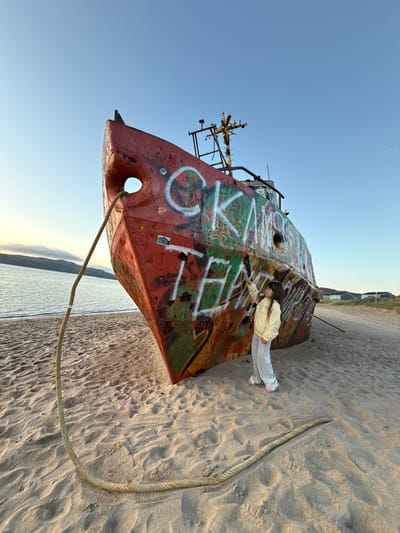 An abandoned, graffitied red ship rests on a sandy beach in Teriberka, Russia, with the sea and distant hills in the background. A young girl wearing a light yellow jacket and grey sweatpants stands beside the ship, reaching out to touch the graffiti. A thick rope extends from the ship's bow and lies on the sand. The sky is clear and blue, indicating it is daytime. Buildings with green roofs are visible in the distance.