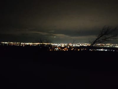 A panoramic nighttime view of the city of Balsha, Bulgaria, is depicted from an elevated, unlit vantage point. The lower half of the frame is entirely dark, representing unilluminated foreground terrain. Above this, a wide, horizontal band of blurred, artificial lights illuminates the distant city, displaying predominant hues of white, yellow, and orange. Silhouetted against the city lights and dark sky, leafless tree branches are visible. A distinct, prominent branch extends from the lower right quadrant upwards and towards the left. The upper portion of the image consists of a dark, overcast sky with discernible cloud formations. A single, faint, bright point of light, possibly a celestial body, is visible in the upper-central area of the sky. No individuals, specific actions, or interactions are discernible.