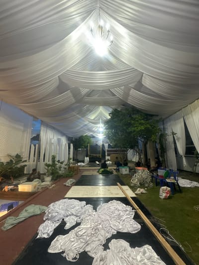 The image depicts the interior of a large white tent structure, likely set up for an event, situated in Langa, Indonesia. The tent is adorned with draped white fabric that forms a vaulted ceiling, illuminated by a central chandelier. In the foreground, several piles of white fabric are spread out on a black and white checkered surface, suggesting they are being prepared or stored. Several individuals are visible in the mid-ground, some sitting and interacting, while others appear to be working or resting. Various objects are scattered around the scene, including plastic containers, chairs, plants in pots, and what appears to be a delivery truck in the background. The overall environment suggests an outdoor or semi-outdoor setting, with glimpses of trees and foliage. The lighting indicates it is either dusk or nighttime.
