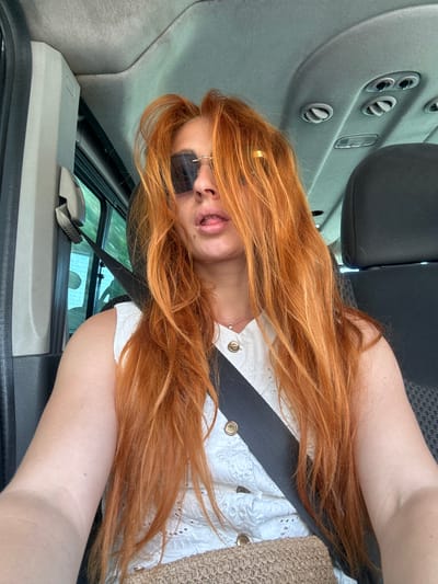 The media file is a selfie of a woman with long, reddish-blonde hair. She is wearing sunglasses and a white top with decorative eyelet detailing and buttons. A straw bag is visible on her right side. The woman appears to be sitting in the passenger seat of a car, indicated by the car's interior visible in the background, including air vents and upholstery. The angle of the photo suggests she is leaning back and to her side. Her expression is playful, with her tongue slightly sticking out. The lighting suggests it is daytime, likely sunny. The location context provided is Rijeka Reževići, Montenegro, which might imply a warm climate and coastal scenery, though these are not directly visible in the frame. No visible text is present in the media file.