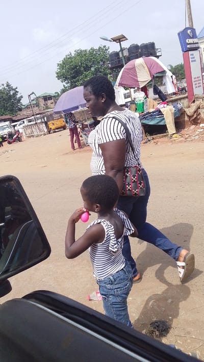 An adult female and a young girl walk along a reddish-brown unpaved road in Igbo Ukwu, Nigeria, under an overcast sky. The female, with dark short hair, wears a white and black striped short-sleeved top, dark blue jeans, and white sandals on her left foot, with a patterned black and pink shoulder bag on her right side. The girl, positioned slightly in front and to the left of the female, wears a white and black striped top with ruffled shoulders, blue jeans, and has a small earring in her left ear. She holds a pink spherical object to her mouth with her right hand.

The scene is viewed from the perspective of a vehicle, with a black car mirror and frame visible in the immediate foreground on the left. In the mid-ground, a vibrant street market environment unfolds. On the left, multiple white vans or buses are parked alongside a yellow auto-rickshaw and a red scooter. A purple market umbrella is open on the left. On the right, a maroon and white striped market umbrella covers displayed clothes. Behind this, a tall utility pole structure supports solar panels and multiple black water tanks. Adjacent to this, a blue and red sign for "OPTIMUM COMPANY OIL" advertises "FUEL KERO DIESEL" and "MAX LUBRICATION." Buildings with green roofs are visible in the distant background, interspersed with trees. Debris, including a plastic bottle and a dark clump of material, lies on the unpaved road surface.