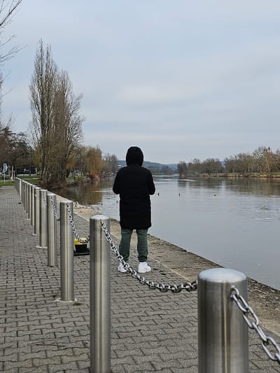 The image depicts an outdoor scene in Bech-Kleinmacher, Luxembourg, along a river promenade under an overcast sky. A single individual stands with their back to the camera on a paved path of grey interlocking bricks, facing a calm, grey-brown river. The individual is dressed in a black hooded long coat, dark green trousers, and white athletic shoes. A barrier of silver-colored cylindrical metal bollards connected by metal chains runs along the edge of the promenade, separating it from the riverbank. A black mooring cleat with a yellow top is visible at the river's edge, behind the barrier. Several waterfowl are present on the water's surface in the midground. The far bank of the river is lined with mostly bare deciduous trees, extending to the horizon where faint hills or structures are discernible under a uniformly light grey sky.