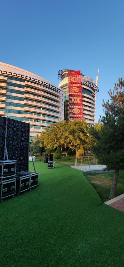 The image depicts a modern building with a distinctive facade featuring curved lines and alternating bands of windows and white panels. A prominent red and gold tapestry or banner is draped across the upper section of the building, adding a cultural or decorative element. The building is situated outdoors, with a lush green lawn area in the foreground and to the right. Trees, some with visible pine needles, are scattered around the area, with sunlight filtering through their leaves, suggesting it is daytime.

In the foreground, several black cases, likely containing audio or lighting equipment, are stacked and arranged on the lawn. Some of these cases appear to be part of a larger setup, possibly for an outdoor event. The sky is a clear, vibrant blue, indicating fair weather. The perspective of the image is from ground level, looking upwards towards the building and trees, and also to the side, encompassing the lawn and equipment. The overall impression is one of an event setup in a modern, landscaped urban environment. The location context of Türkmenbaşy, Turkmenistan, suggests a possible cultural or official event, given the architectural style and the prominent tapestry. No visible text is present in the image.