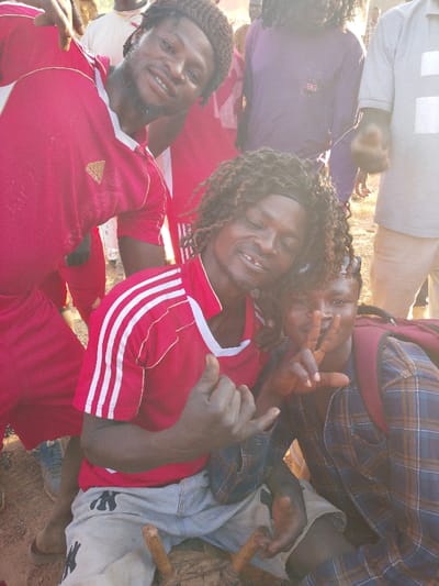 The image captures an outdoor gathering in Vodni, Nigeria, under bright natural light. In the immediate foreground, three individuals pose. To the left, a man with a dark knitted cap, braided hair, and a beard wears a red Adidas shirt with white stripes; he smiles directly at the camera with his right hand partially raised. Central in the foreground, another man with curly hair and a dark headband, wearing a matching red Adidas shirt and light grey shorts with a dark "NY" logo, sits on a low wooden object. He grins broadly, revealing gaps in his upper teeth, and makes a 'shaka' hand gesture with his left hand. Partially visible behind him to the right, an individual wearing a blue and brown plaid shirt and a red backpack holds up their right hand in a 'peace' sign. The ground is dry, reddish-brown dirt. Multiple additional individuals are present in the blurred background, including one in a white top, one in a purple top, and another in a grey t-shirt.