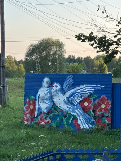 This image depicts an outdoor scene in the city of Lysaya Gora, Russia, featuring a colorful mosaic artwork on a blue wooden fence. The mosaic, made of bottle caps, depicts two white doves with blue accents on their wings, facing each other, with red beaks touching. They are positioned above a cluster of red flowers with green centers and stems, and some green foliage. The artwork is located on a blue, horizontally planked fence. To the left of the fence, a wooden utility pole with wires running overhead is visible. The background consists of lush green grass, scattered small white wildflowers, and a dense line of trees under a clear, pale sky. The overall impression is one of folk art decoration in a rural or semi-rural setting. There are no people visible in the image.