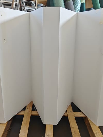 The image shows a large, white, folded structure. It appears to be made of a rigid material, possibly cardboard or a similar composite, and is arranged in a zigzag or accordion-like pattern with several angled panels. The structure is supported from underneath by wooden planks, suggesting it might be a temporary installation or a prop.
In the background, behind the white structure, hints of a storage or workshop environment are visible. There are metal ladders leaning against a wall, and rolls of what appear to be carpet or fabric are stacked up, some wrapped in plastic. The lighting suggests it is daytime, likely indoors, as there are no strong shadows or indications of natural outdoor light.
The focus of the image is the white structure itself. There are no people visible in the frame. The setting is likely a commercial or industrial space, possibly a warehouse, a workshop, or a storage area for materials and equipment. The overall impression is one of a behind-the-scenes or preparation area, rather than a finished or public space. The white surface of the structure has some minor imperfections, such as smudges and two small dark dots on the leftmost panel. A small piece of white tape is affixed to the rightmost panel. The wooden supports are rough-hewn planks, some showing nails or screws.
