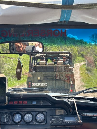 A vehicle in Haghartsin, Armenia follows another vehicle on a rural road. The interior of the vehicle includes a rear-view mirror with a reflection of the driver, a radio, an Areon air freshener, dashboard gauges, and a curtain. The license plate of the vehicle ahead reads "37 LF 690." The other vehicle is an open-top vehicle with people in it. The sky is visible through the front windshield.
