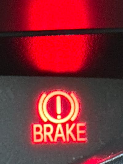 The image shows a close-up of a vehicle's dashboard warning light illuminated in red. The warning light displays a symbol of a circle with an exclamation mark inside, and the word "BRAKE" is written below it. The background is dark, with a blurred red light above the warning indicator. There are no people, animals, or recognizable landmarks of Lynnwood, United States visible in the image. The scene is limited to the illuminated dashboard indicator.