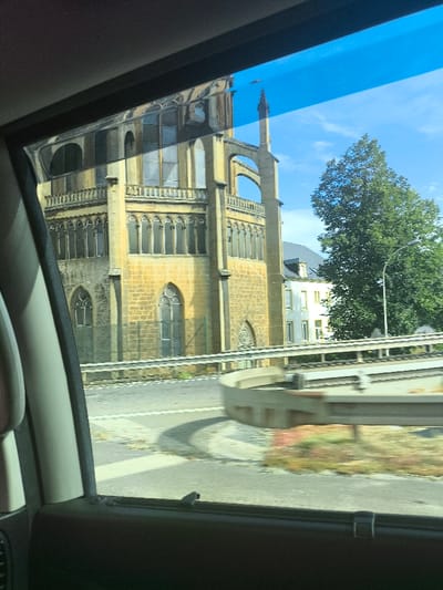 A large, yellowish, multi-story building with arched windows and decorative elements is seen through a car window. The building appears to be made of stone or brick and has a tower-like structure with an arch. A road with a guardrail runs in front of the building. A green tree and a white building are visible in the background. The sky is blue with some clouds. The image is taken from inside a car, with the car's interior visible in the foreground.