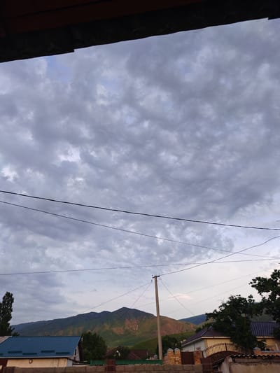 The image depicts an overcast sky with heavy, textured clouds. In the foreground, there is a dark wooden overhang, possibly part of a building. Several power lines are strung across the sky, creating a linear pattern against the clouds. Below the power lines, a residential area is visible, with houses featuring various roof colors, including blue and dark brown. One house has a light-colored facade and a decorative metal gate. Further in the background, rolling green hills and mountains rise, some with patches of sunlight illuminating their slopes, suggesting a dramatic lighting contrast. A lone utility pole stands prominently in the middle ground. The scene appears to be in a semi-rural or suburban setting, consistent with the outskirts of a city like Кашка-Суу, Kyrgyzstan, given the presence of residential buildings, open land, and mountainous terrain. No people are visible in the frame.