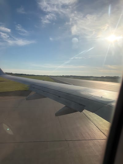 The image depicts a view from an airplane window. The plane appears to be on a runway, with a wing visible in the foreground. The sky is visible in the upper left, with the sun shining brightly. Adjacent to the runway is an expanse of greenery, presumably grassy fields, stretching towards the horizon. In the background is a tree line. No people are visible, and the location within Crokhovo, Russia is unidentifiable from the image.
