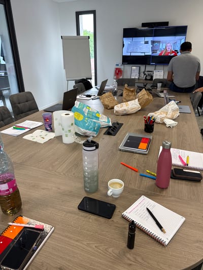 The image depicts a meeting room with a large wooden table. On the table are various items including laptops, notebooks, pens, paper rolls, a water bottle, coffee cup, and food packaging. A large screen is mounted on the wall, displaying a video conference with two participants. A man is seated on the edge of the table, facing the screen. A whiteboard is visible in the background. The room has large windows, suggesting it is daytime. The scene suggests a business meeting or presentation is taking place.