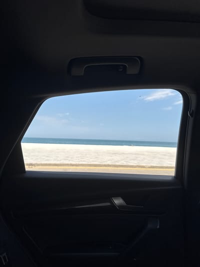 The view from inside a vehicle frames a scene of Boca del Río, Mexico. Outside the window, the sky is blue with scattered clouds, meeting the ocean along the horizon. A sandy beach stretches to the right, marked by a yellow line. The interior of the vehicle is dark, with the window surrounded by black upholstery and car fixtures like a grab handle and trim.