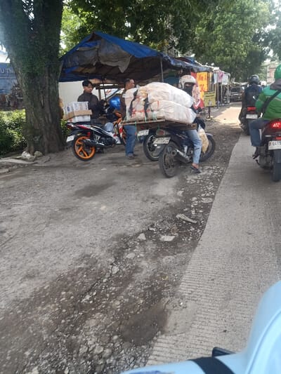 A street scene shows several motorbikes parked or moving along a road. One is heavily laden with large white bags stacked on a wooden pallet. Two men stand near the bikes. Other motorbikes, including one with a green-shirted rider, are further down the road. A vendor stall with a blue tarp is next to a large tree. Additional stalls line the street beyond. The road surface is uneven and cracked in places.