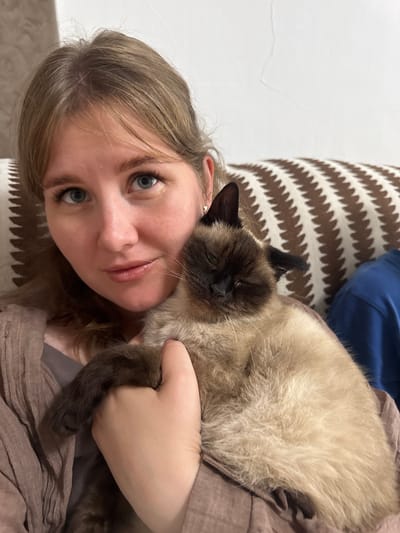 A close-up shot shows a young woman with blonde hair and blue eyes holding a light brown Siamese cat. The woman is wearing a light brown cardigan over a grey top. The cat is nestled in her arms, with its eyes closed and its face partially obscured. The background features a couch with a striped pattern in brown and cream, and a white wall. The scene appears to be indoors. The location of Буб, Russia, is not evident in the image.