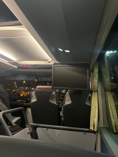 The image displays the interior of a public transport vehicle, likely a bus or train, during nighttime. The perspective looks towards the rear or middle section of the passenger cabin.
Multiple rows of dark, textured seats with prominent light-colored (white or light grey) armrest trims are visible. Above the seating area, light-colored curtains are neatly tied back, revealing large windows. Through these windows, blurred lights from an urban or industrial area are visible against the dark night sky, indicating it is nighttime.
Overhead, white or light grey panels house several small, circular lights and a long, linear light strip that illuminates the interior. A flat-screen monitor, currently displaying a dark screen, is mounted on a support structure, presumably for passenger entertainment or information.
A small red digital display is embedded in one of the overhead panels, showing the numbers "16.3", which could represent temperature or time. Reflected prominently in the windows or the monitor's surface, in reversed and a distinctive gothic-style script, is the word "Donnerstag" (German for Thursday), suggesting a timetable, destination, or other informational text.
The setting is a modern, well-lit vehicle interior, implying travel. No specific activity is clearly depicted, though the arrangement suggests passengers are seated. A faint outline of a person's head is barely discernible behind one of the seats. The visible German text aligns with the provided location context of Weeze, Germany.